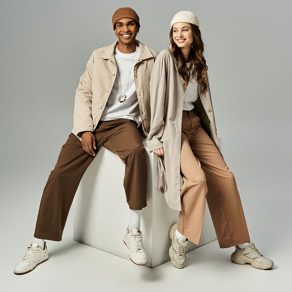 A man and a woman pose together on a white cube in a studio setting, wearing coordinated neutral-toned outfits. The man wears a beige jacket, brown pants, a brown beanie, a white shirt, and white sneakers.